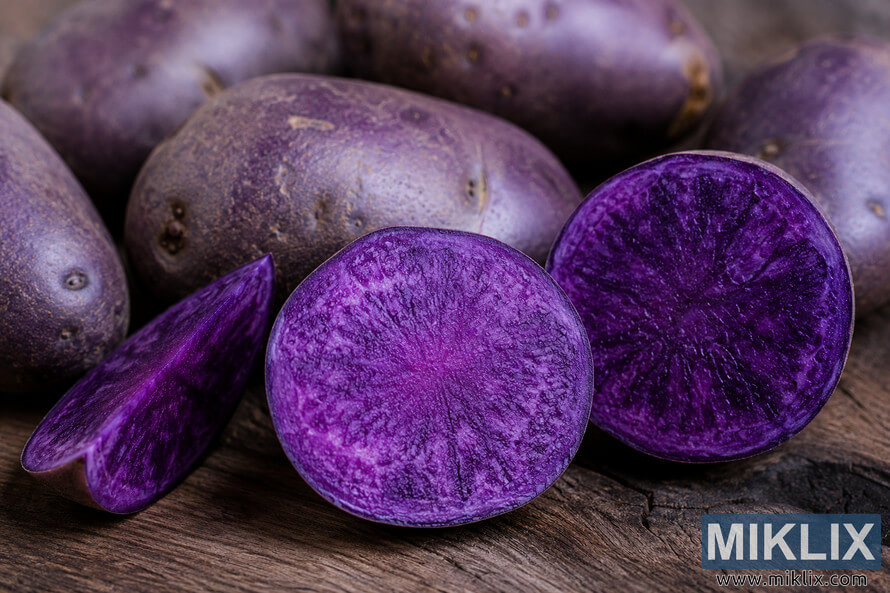 Close-up landscape photograph of whole and freshly cut purple potatoes on a rustic wooden surface, highlighting their vibrant antioxidant-rich violet flesh with detailed natural textures and soft, diffused lighting. Close-up landscape photograph of whole and freshly cut purple potatoes on a rustic wooden surface, highlighting their vibrant antioxidant-rich violet flesh with detailed natural textures and soft, diffused lighting.