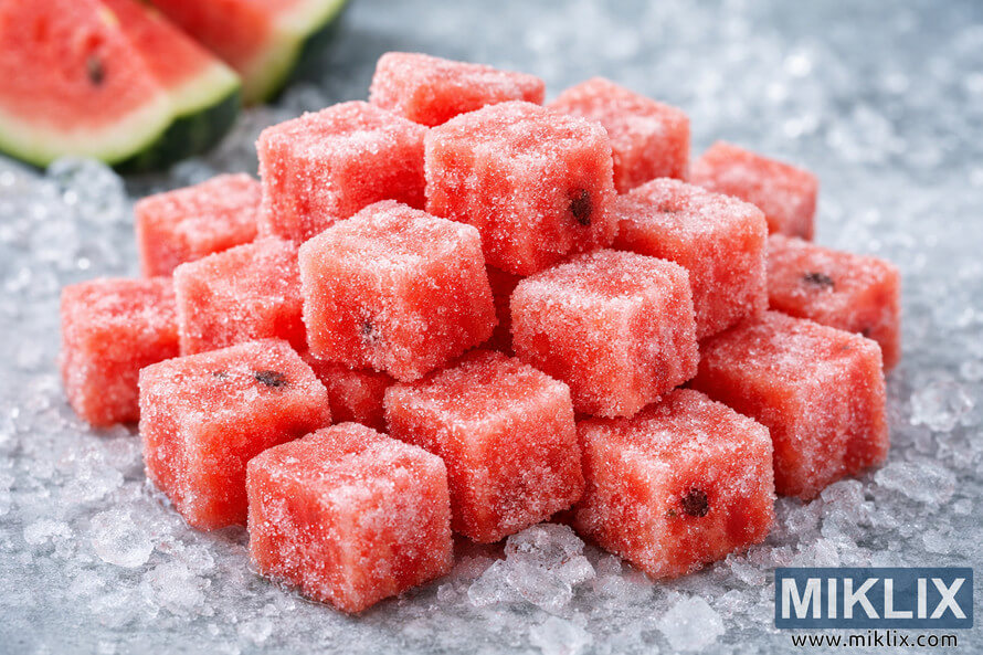 Close-up photograph of a pile of frozen watermelon cubes coated in frosty ice crystals, resting on an icy surface with crushed ice and blurred watermelon slices in the background. Close-up photograph of a pile of frozen watermelon cubes coated in frosty ice crystals, resting on an icy surface with crushed ice and blurred watermelon slices in the background.