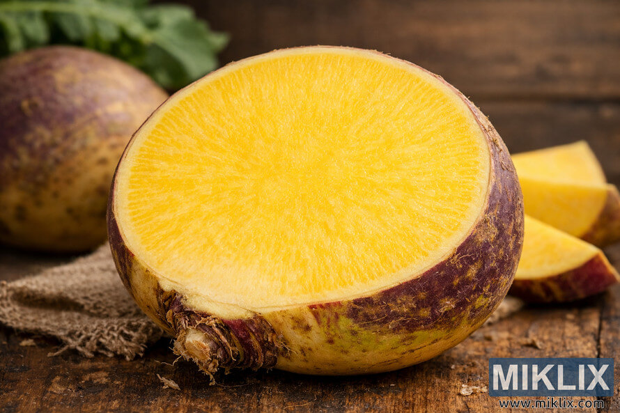 Close-up of a halved rutabaga showing its vibrant yellow flesh and textured purple-brown skin on a rustic wooden surface.