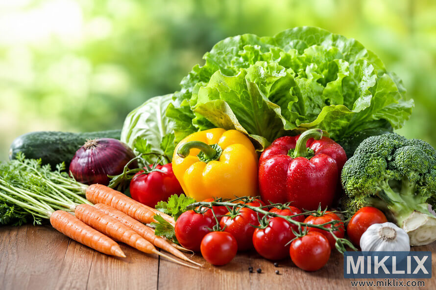 A vibrant assortment of fresh vegetables including crisp green lettuce, red and yellow bell peppers, cherry tomatoes, broccoli, carrots, cucumber, onion, and garlic arranged on a rustic wooden table with a soft green natural background. A vibrant assortment of fresh vegetables including crisp green lettuce, red and yellow bell peppers, cherry tomatoes, broccoli, carrots, cucumber, onion, and garlic arranged on a rustic wooden table with a soft green natural background.
