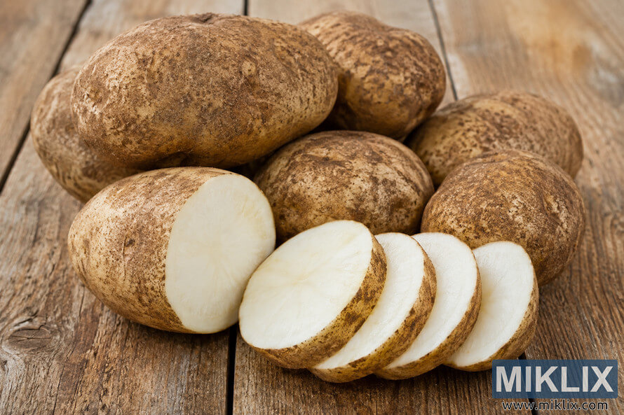 A landscape-oriented close-up photograph of several russet potatoes resting on a rustic wooden table. One potato is cut in half to reveal its pale, starchy interior, while several thick slices are arranged in front. Soft natural lighting highlights the rough brown skins and smooth white flesh against a warm wooden background. A landscape-oriented close-up photograph of several russet potatoes resting on a rustic wooden table. One potato is cut in half to reveal its pale, starchy interior, while several thick slices are arranged in front. Soft natural lighting highlights the rough brown skins and smooth white flesh against a warm wooden background.