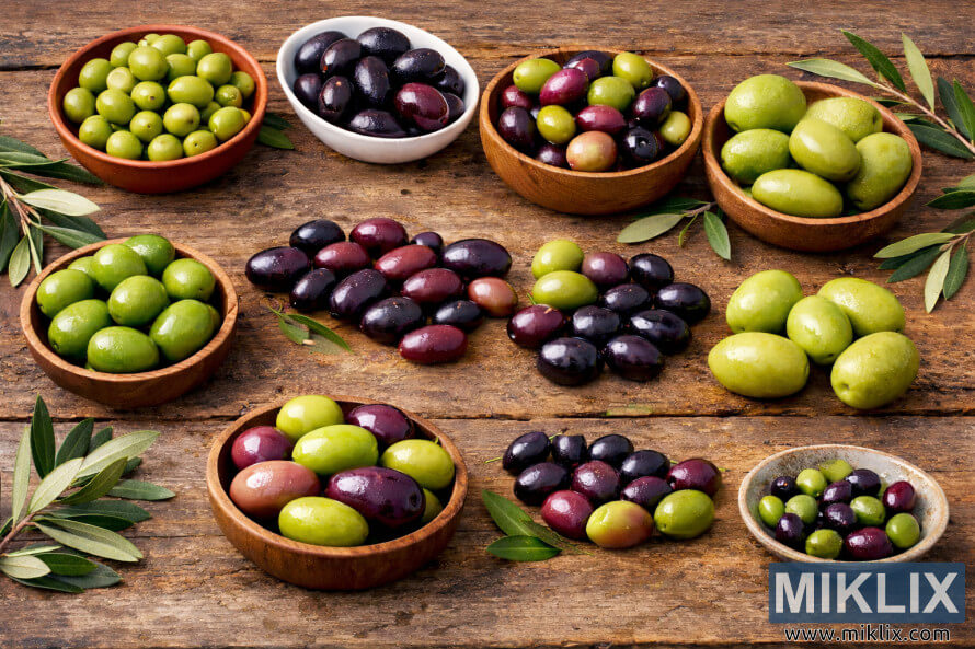 Assorted green, purple, and black olives of different sizes arranged in bowls on a rustic wooden table with olive branches. Assorted green, purple, and black olives of different sizes arranged in bowls on a rustic wooden table with olive branches.