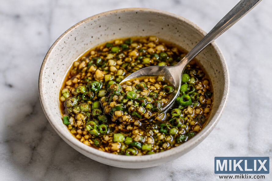 Close-up food photograph of a rustic ceramic bowl filled with glossy ginger scallion sauce on a marble countertop, featuring chopped green onions, minced ginger, and a metal spoon resting inside the bowl.