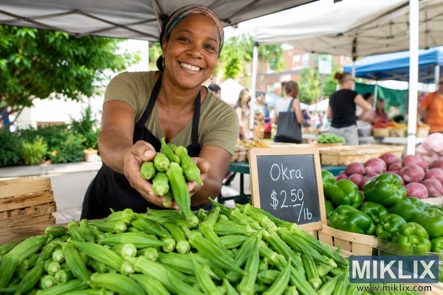 A smiling farmers market vendor extends a handful of fresh green okra toward the camera while standing behind a market stall filled with okra and green peppers beneath white canopy tents. Blurred shoppers and additional produce stalls create a lively outdoor market atmosphere. A smiling farmers market vendor extends a handful of fresh green okra toward the camera while standing behind a market stall filled with okra and green peppers beneath white canopy tents. Blurred shoppers and additional produce stalls create a lively outdoor market atmosphere.