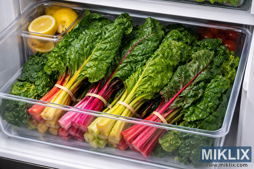 Fresh Swiss chard bundles stored neatly in a refrigerator crisper drawer alongside lemons, broccoli, and cherry tomatoes.
