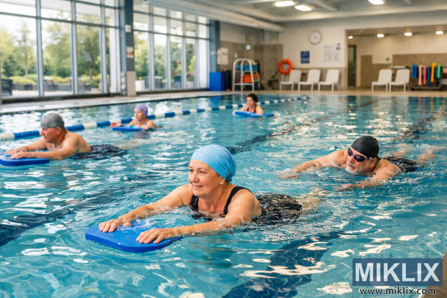 Group of adults using foam kickboards for low-impact exercise in a bright indoor swimming pool with large windows.